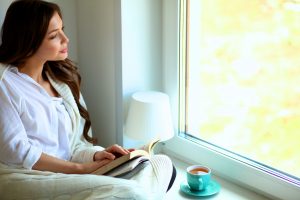 Young woman at home sitting near window relaxing in her living room reading book and drinking coffee or tea .